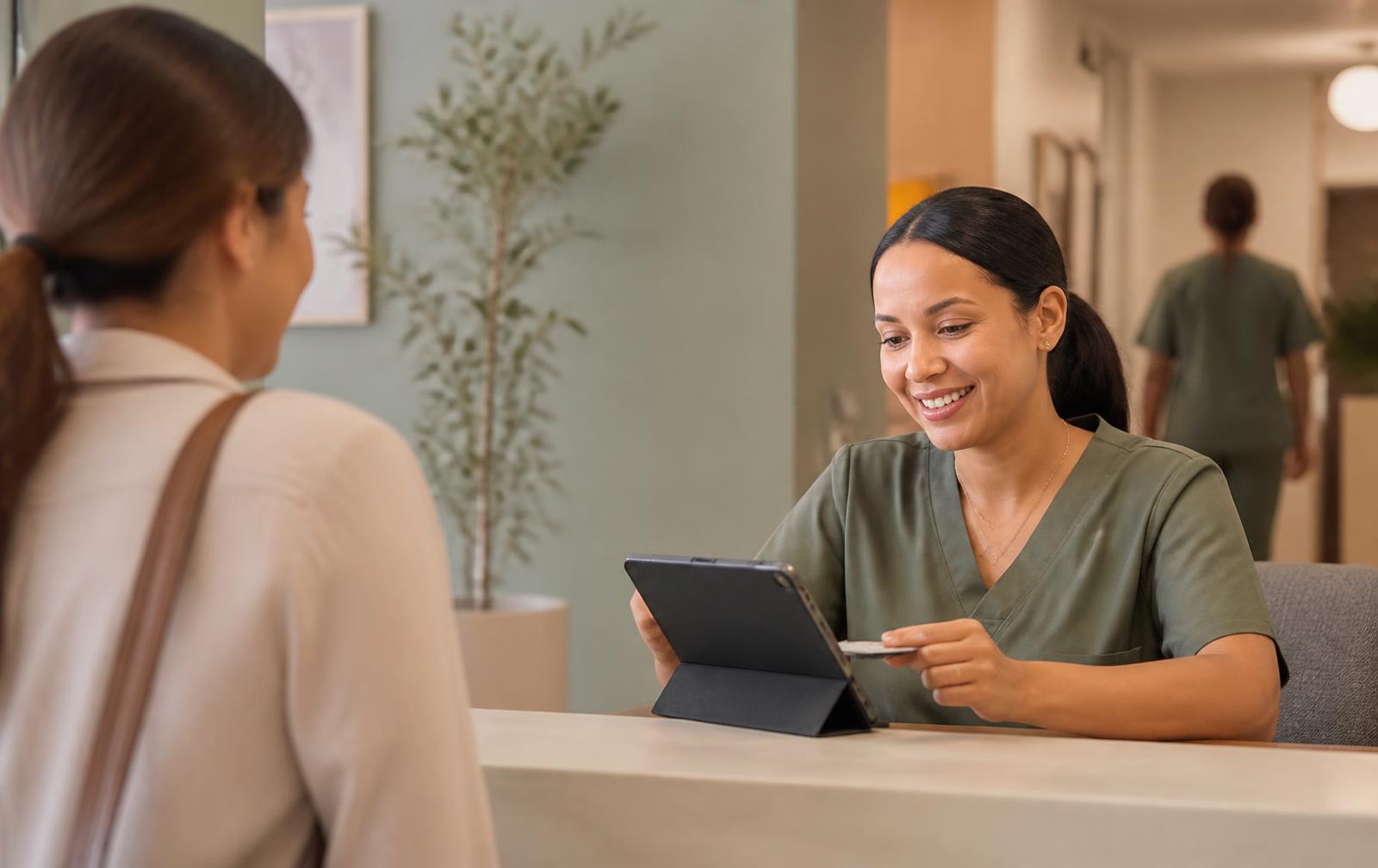 A front-desk staff member verifying a patient's insurance card on a tablet at clinic check-in