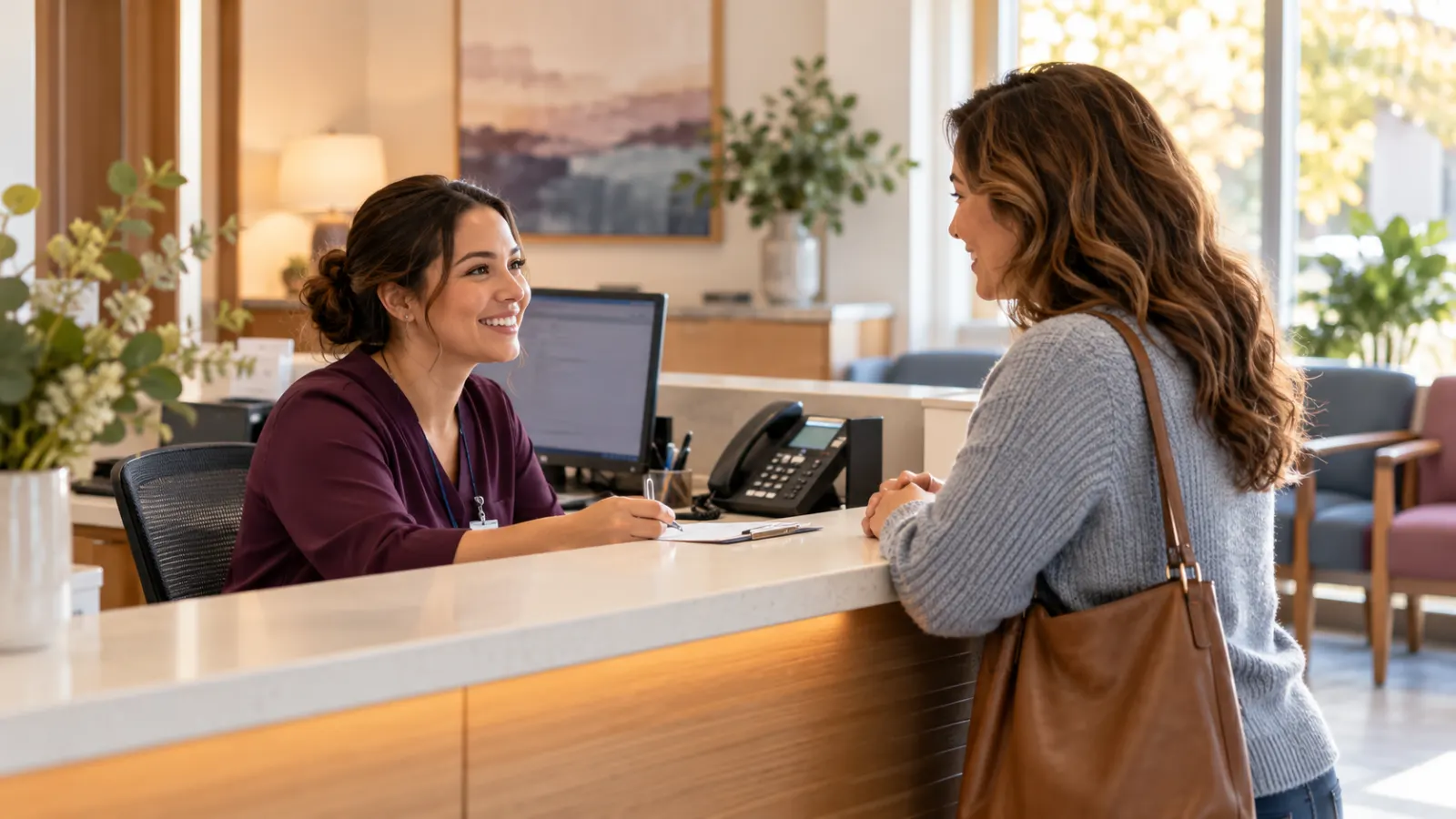 A medical receptionist welcoming a patient beside a desk phone and computer